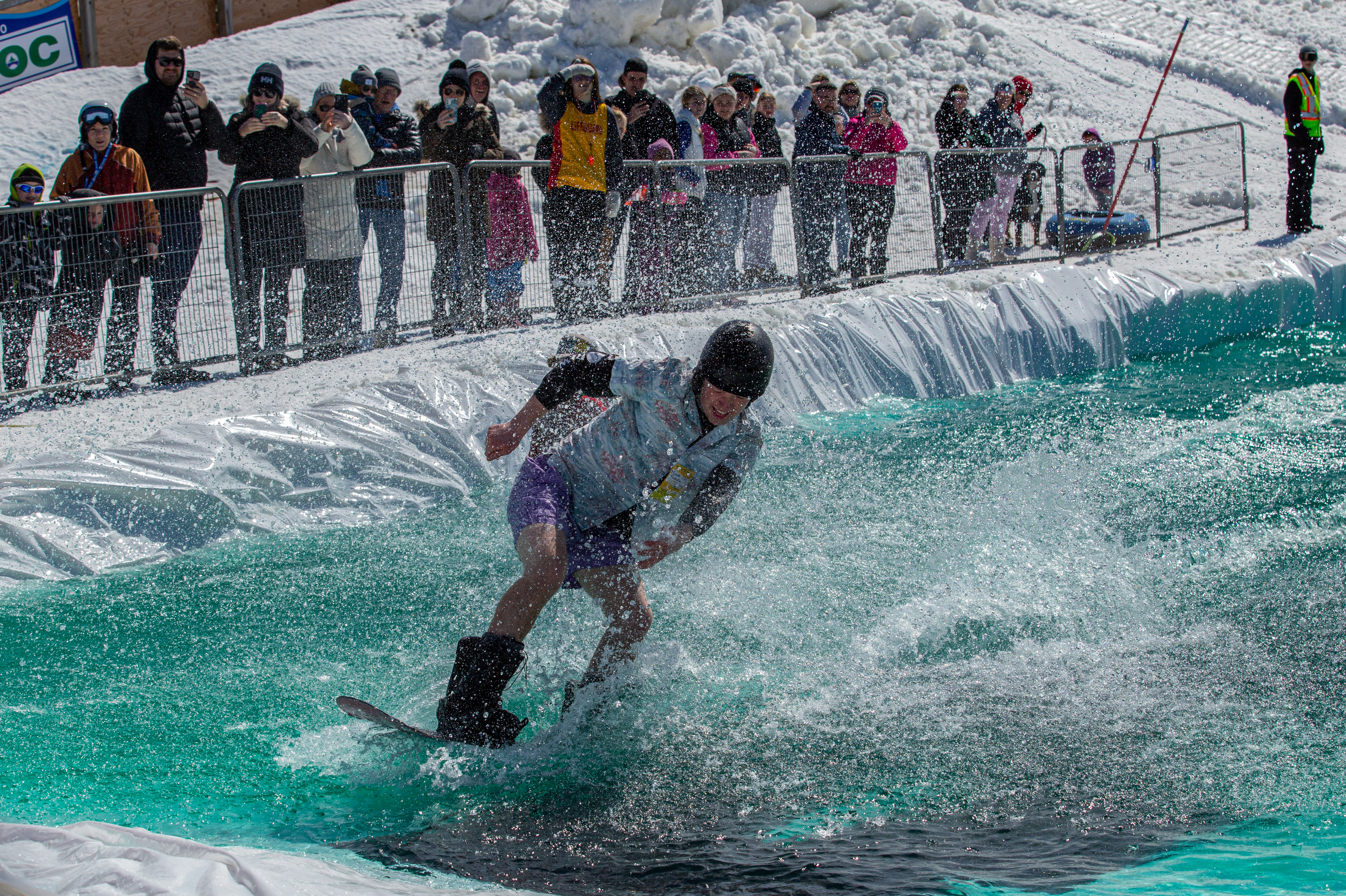 A snowboarder skids across the surface of the water at the Puddle Jump event, with spectators watching from the snowy hillside in the background.