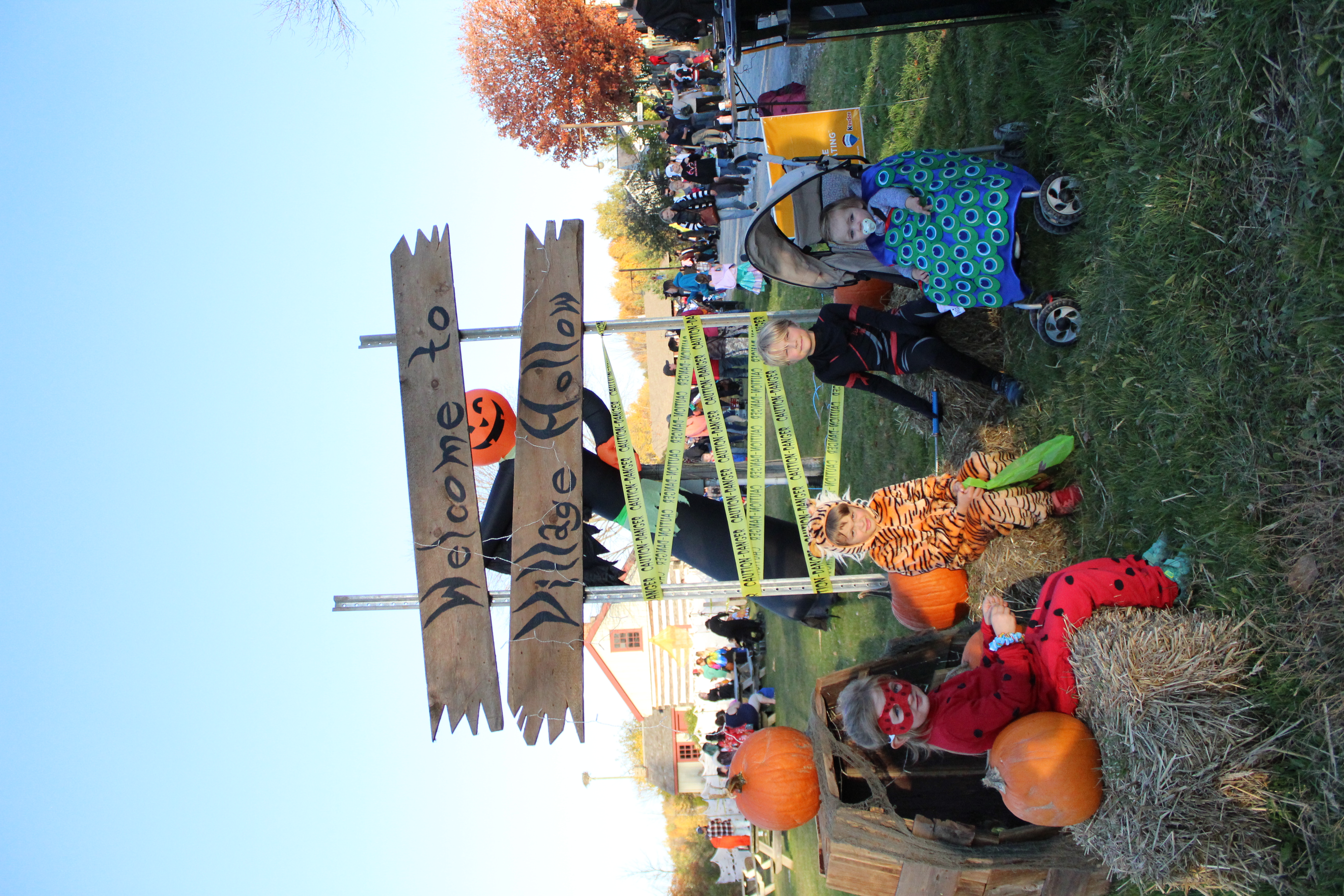 Children in Halloween costumes sit on hay bales in front of a wooden sign that reads “Welcome to Village Hollow.” The scene includes pumpkins, Halloween decorations and a crowd in the background at an outdoor fall event.