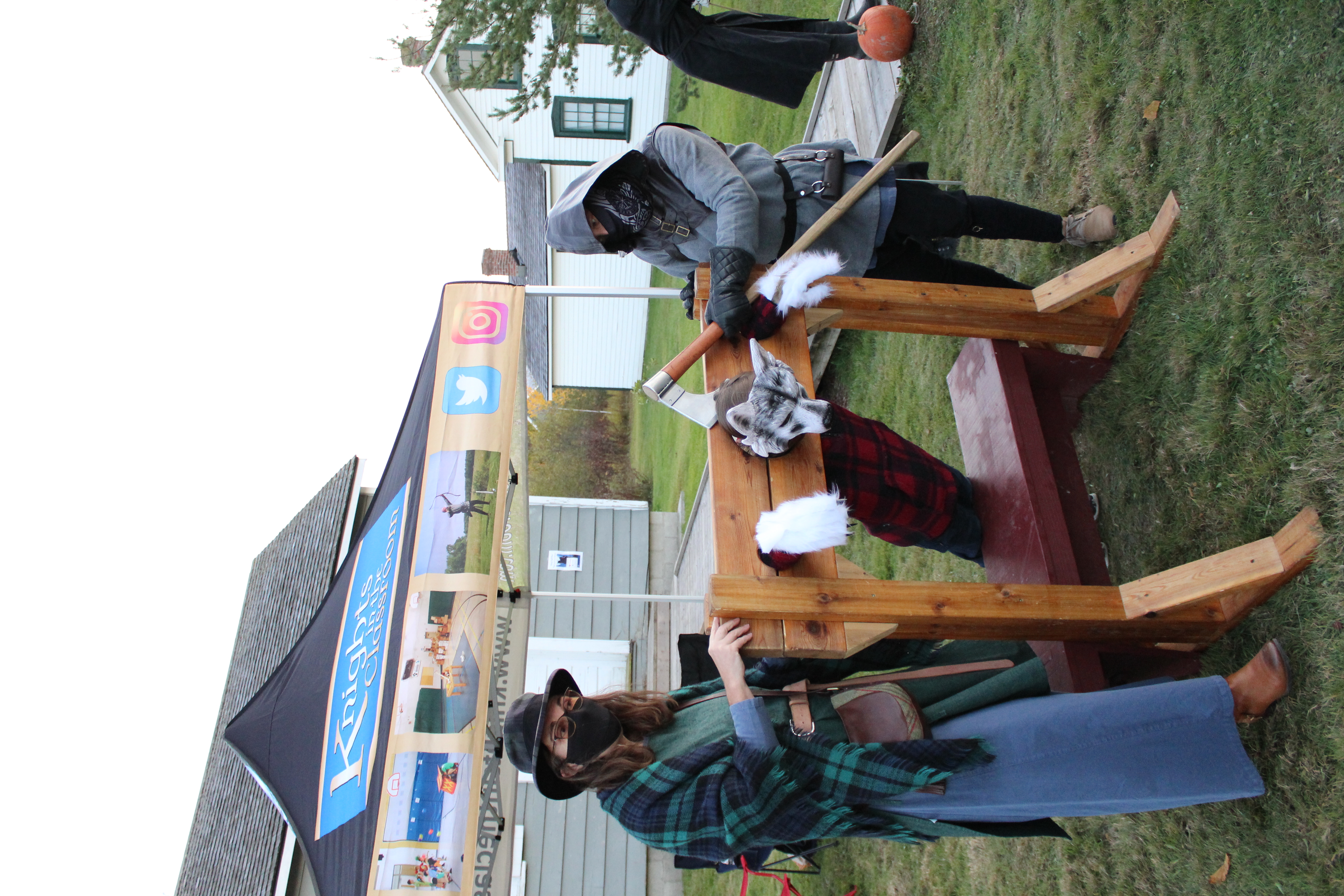 person dressed as a warewolf in wooden stockae with person dressed as an assasin holds axe to their neck and another person dressed as a cowgirl stands beside