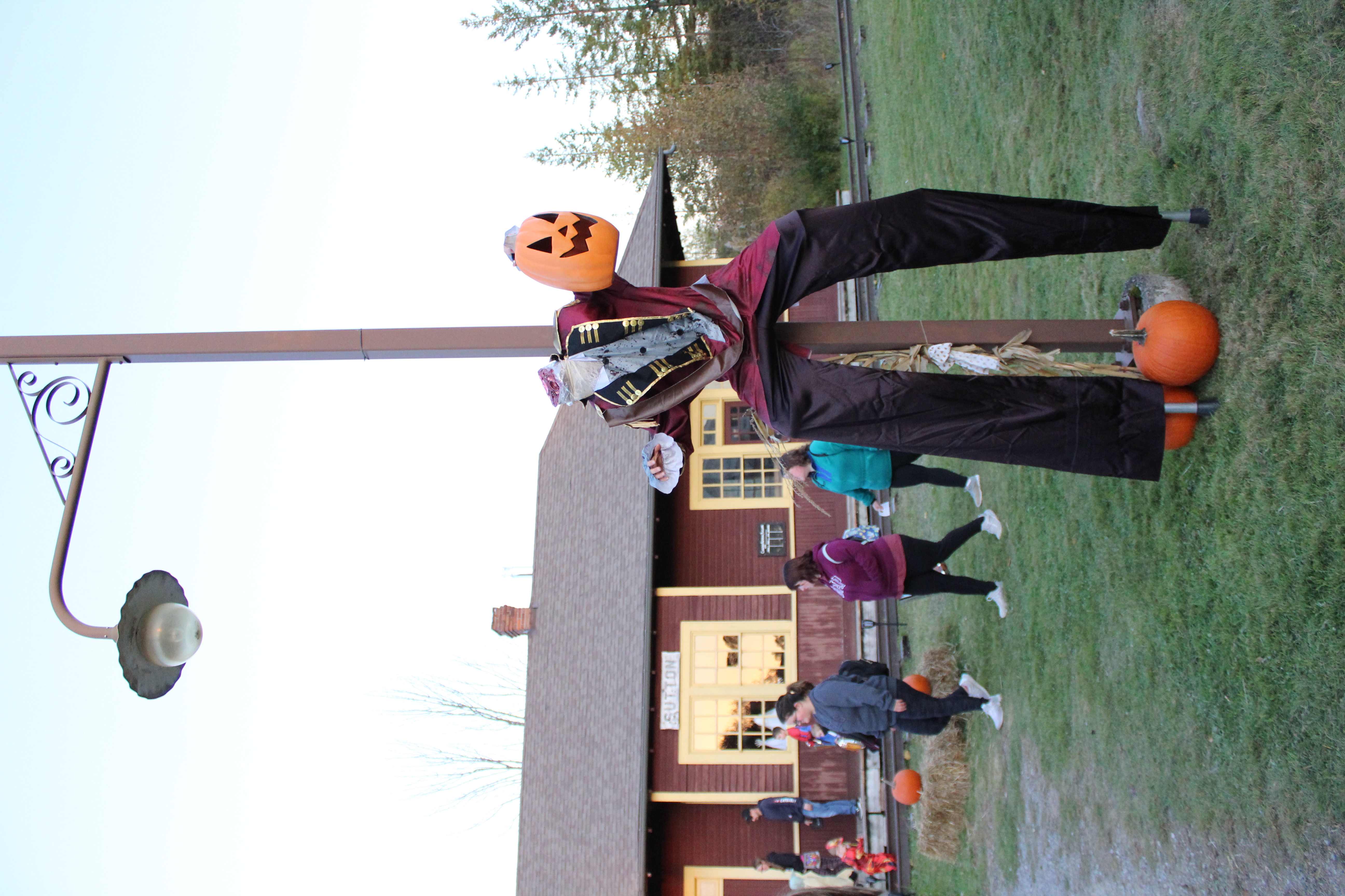 Halloween decoration of headless pumpkin man attached to a lightpost with people walking behind and a fall scene