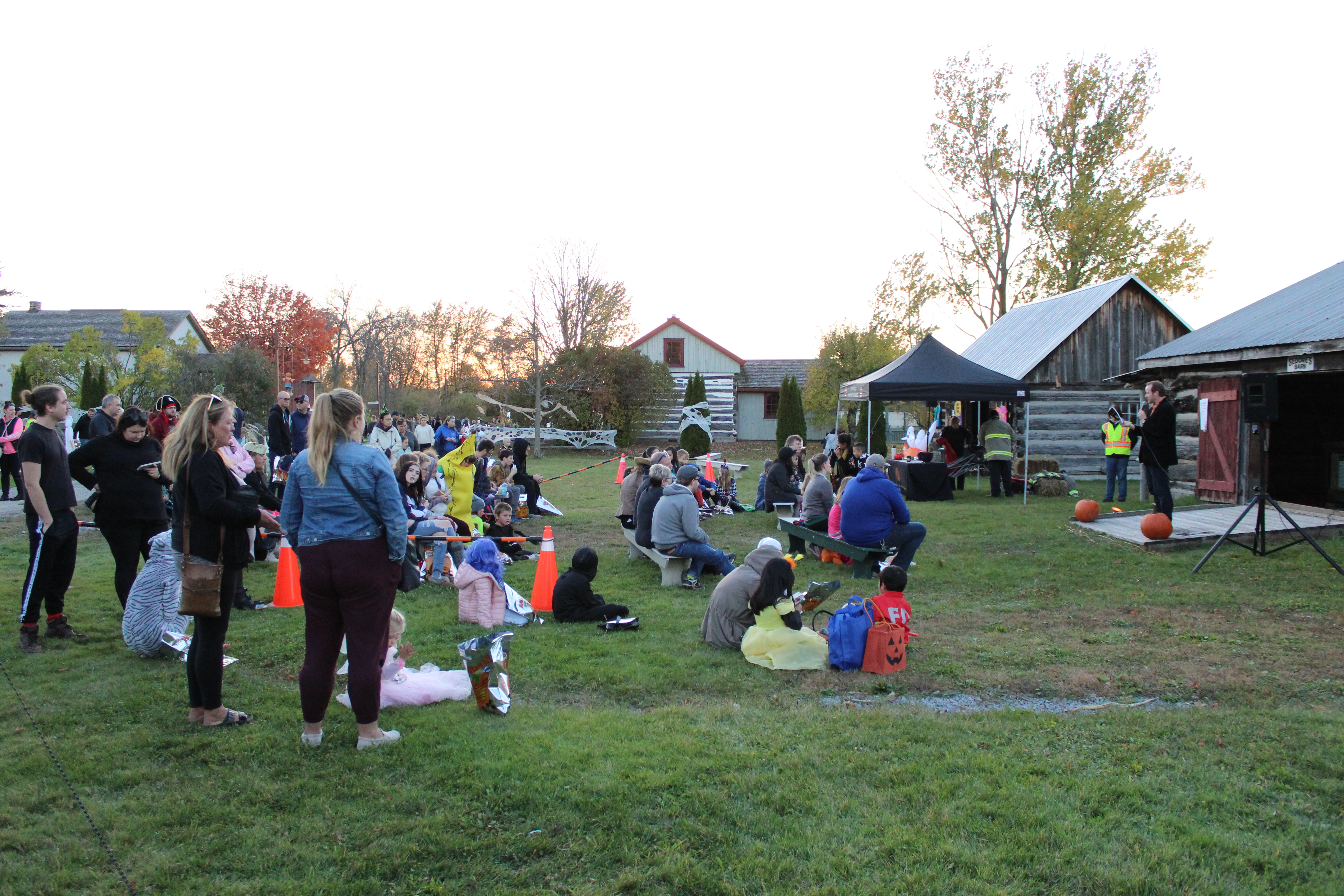 Families and children in Halloween costumes gather on benches and lawn chairs to watch a speaker in front of a historic log cabin. A black tent is set up beside the stage, and the area is decorated with pumpkins and fall-themed displays.