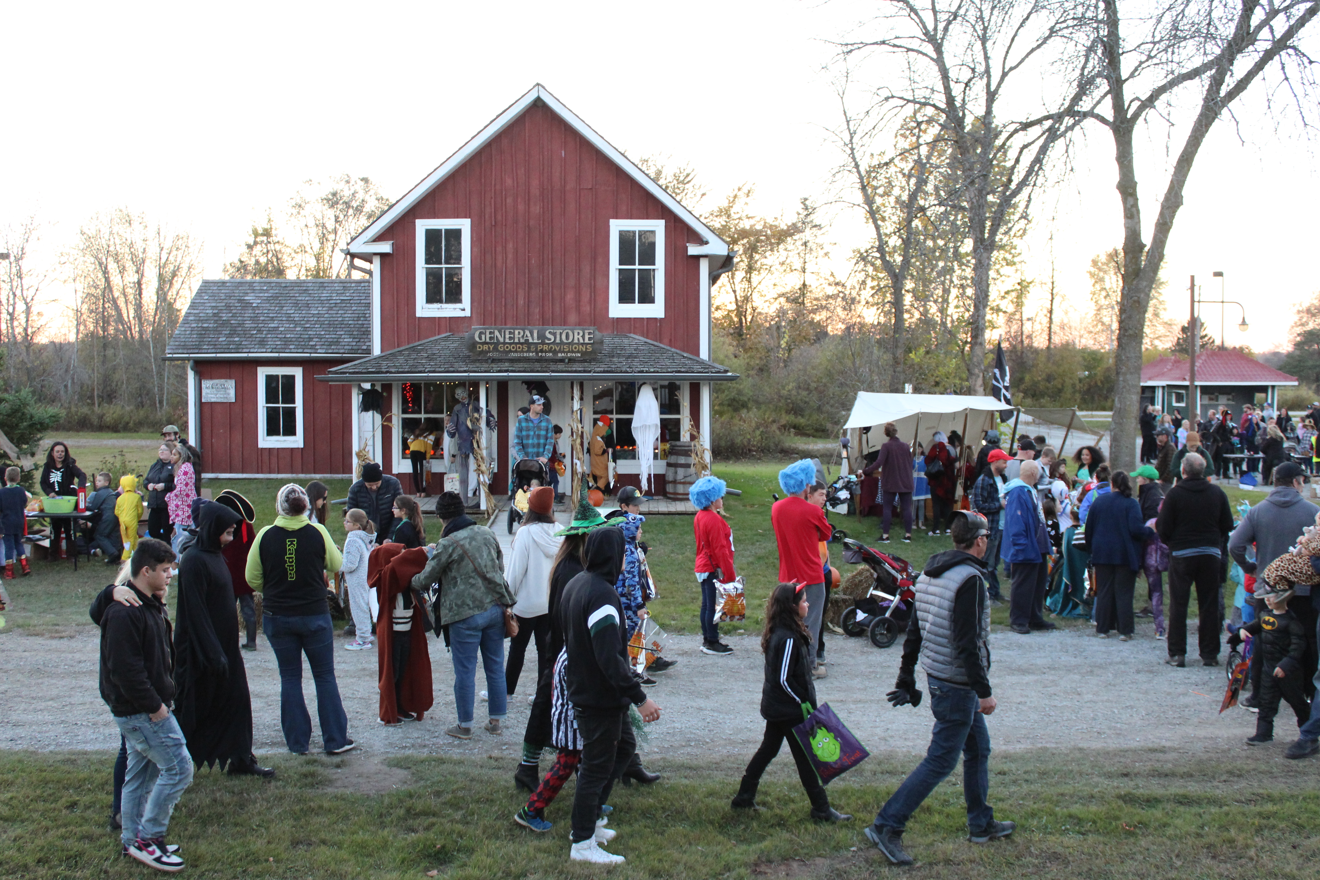 A cheerful family fall event scene: a group of children and adults, many in Halloween costumes like princess dresses and capes, stand and sit on grassy grounds facing a historic log cabin. A speaker addresses them from a small stage beside a black canopy tent, and the gathering is accented with pumpkins, safety cones, and autumn trees in the background.