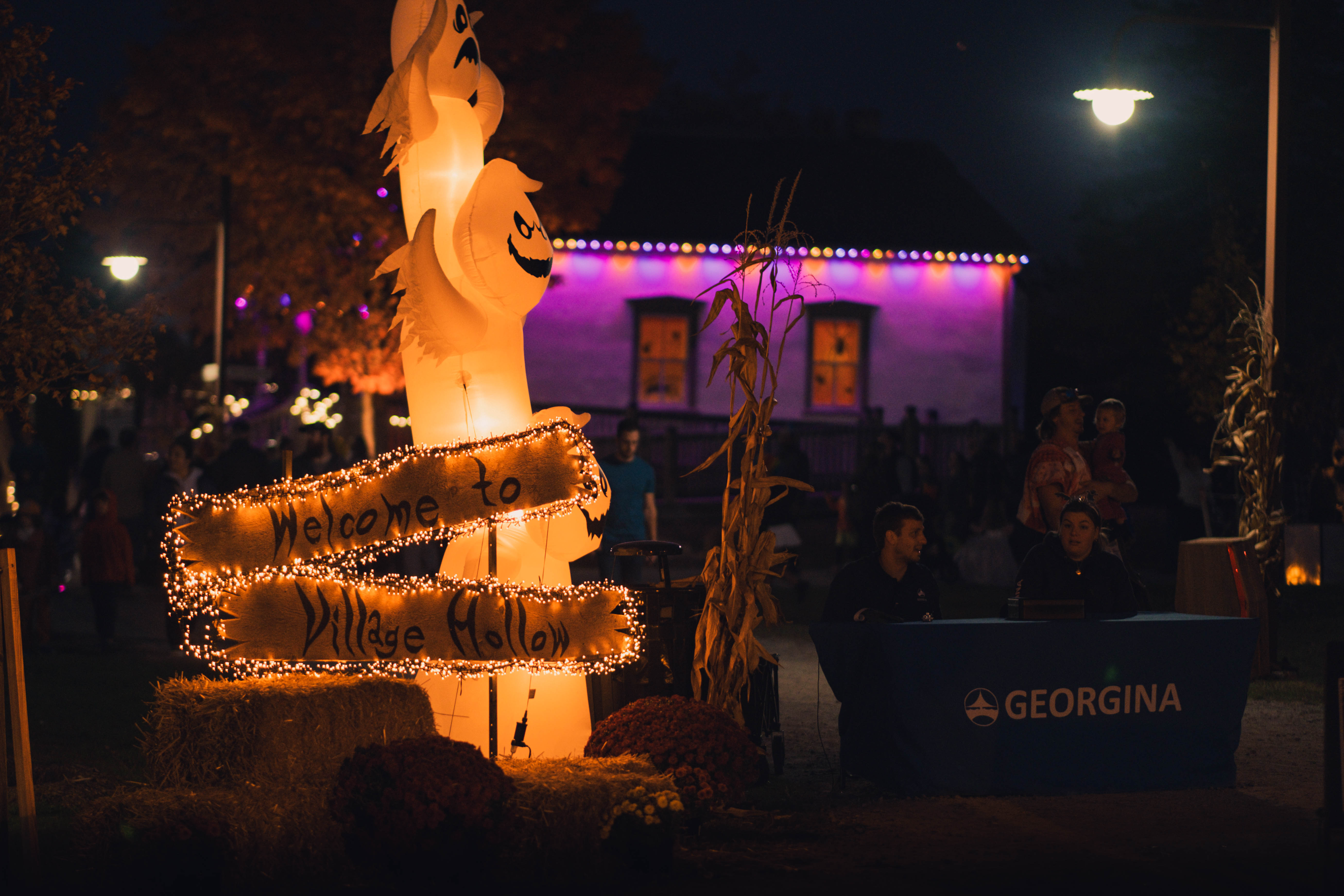 Lit up sign stating welcome to Village Hollow  with table beside with word Georgina and logo of Town of Georgina  on the table. Two people are sitting at the table