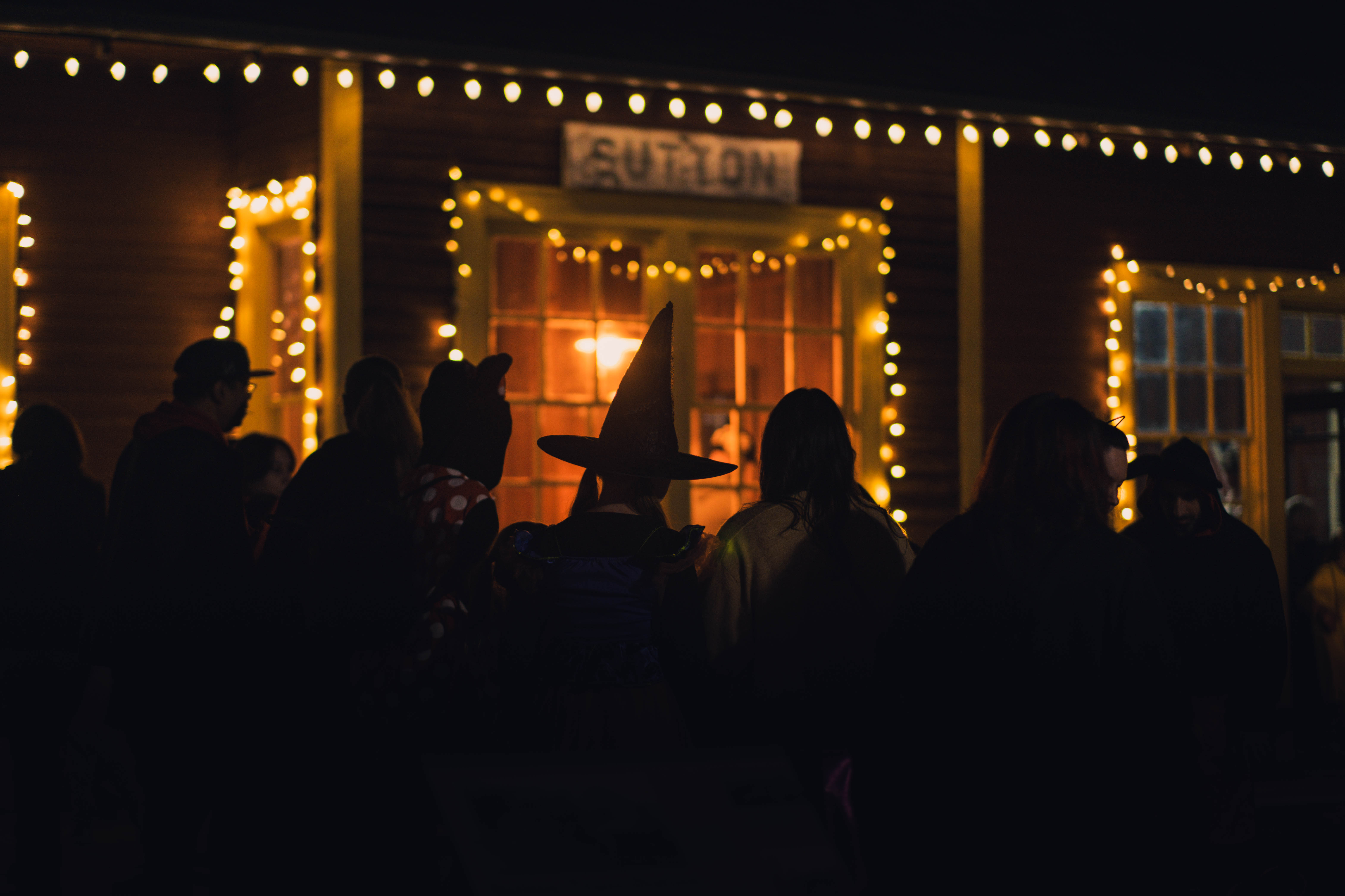 People on shadow with a woman in a witch hat, building behind is lit up with fairy lights around a window with a sign that says sutton