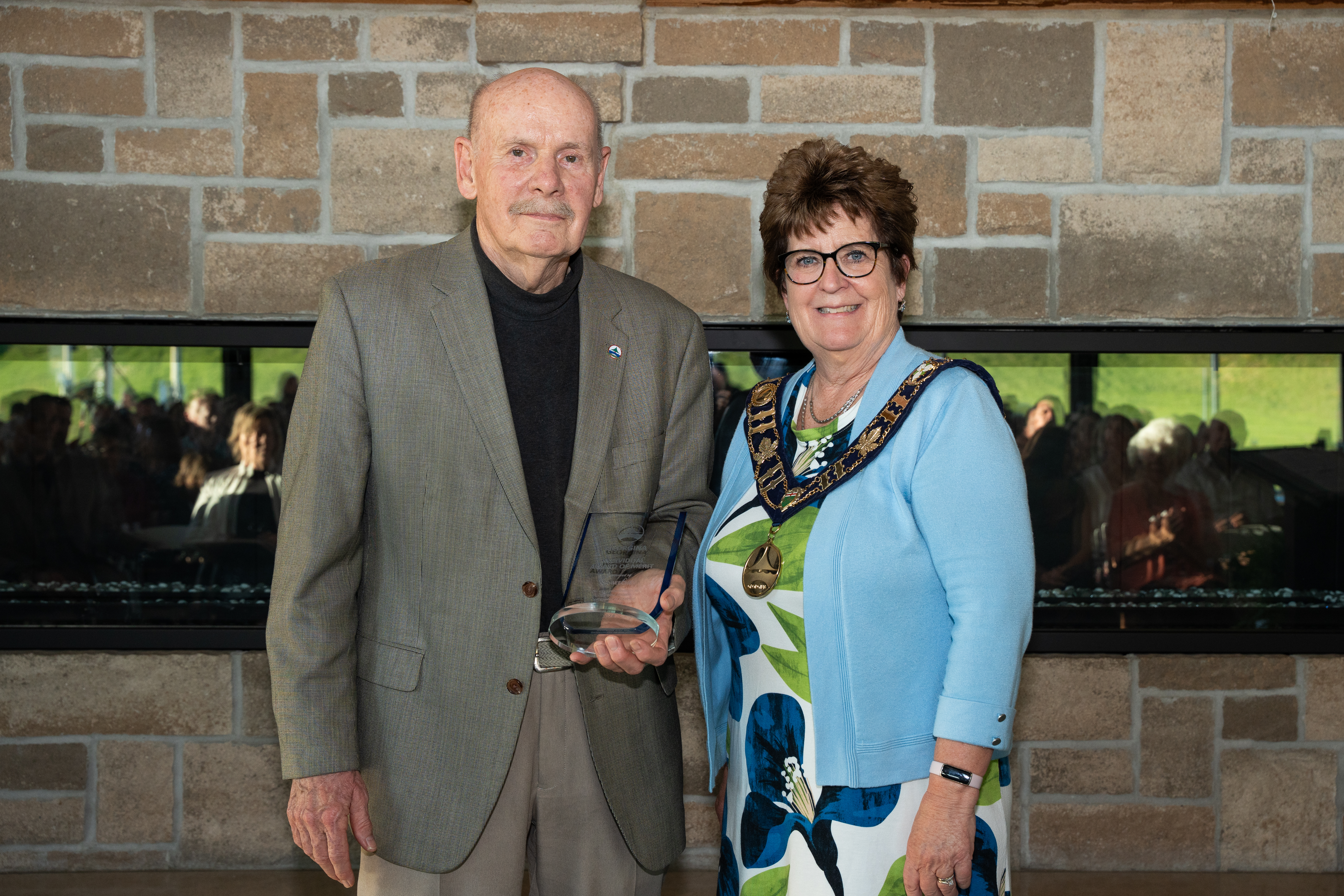 The mayor stands beside an award recipient in front of a brick fireplace, both smiling for the photo.