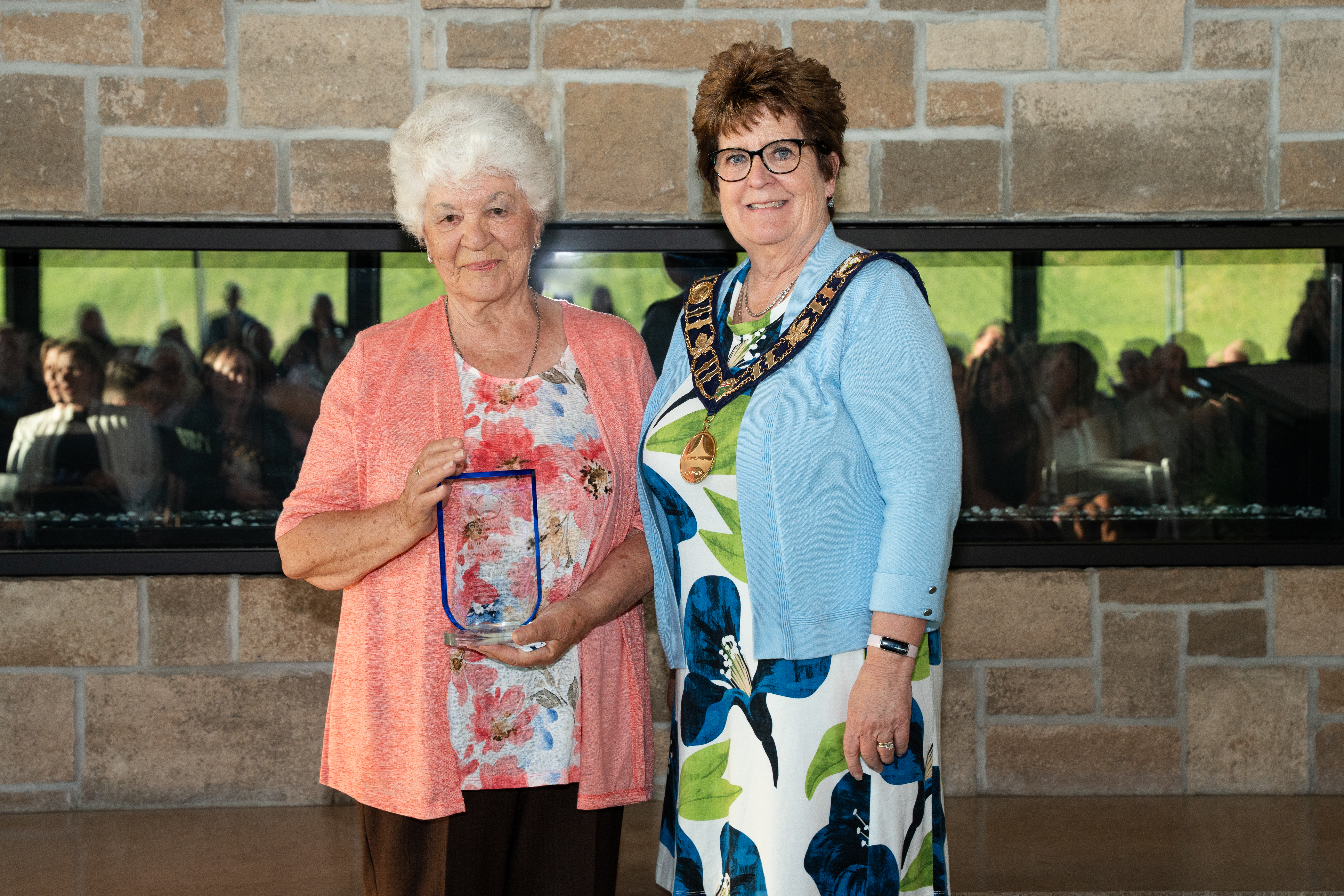 The mayor stands beside an award recipient in front of a brick fireplace, both smiling for the photo.