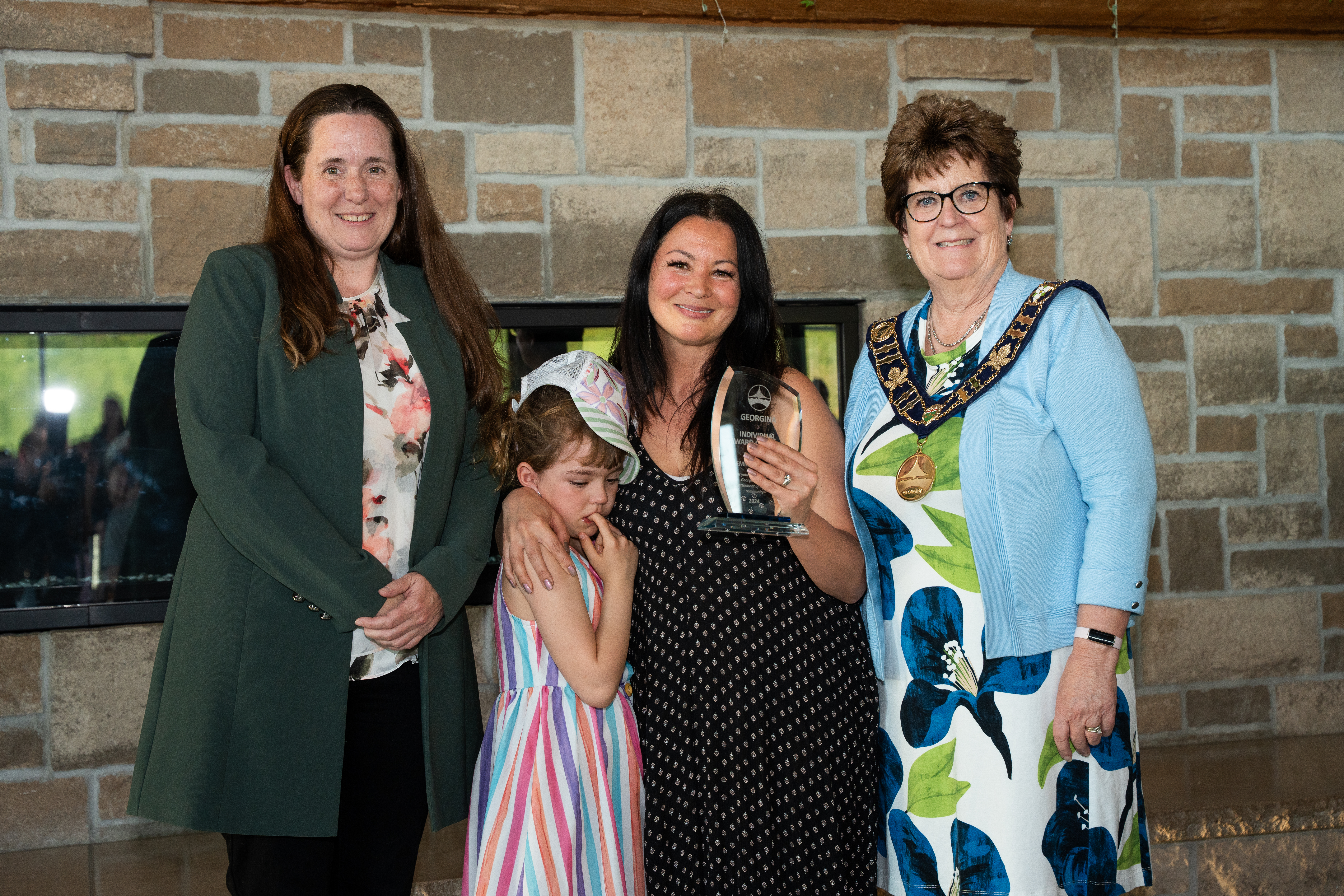 The mayor and Regional Councillor Davidson stand beside an award recipient and a child in front of a brick fireplace, all are smiling for the photo.