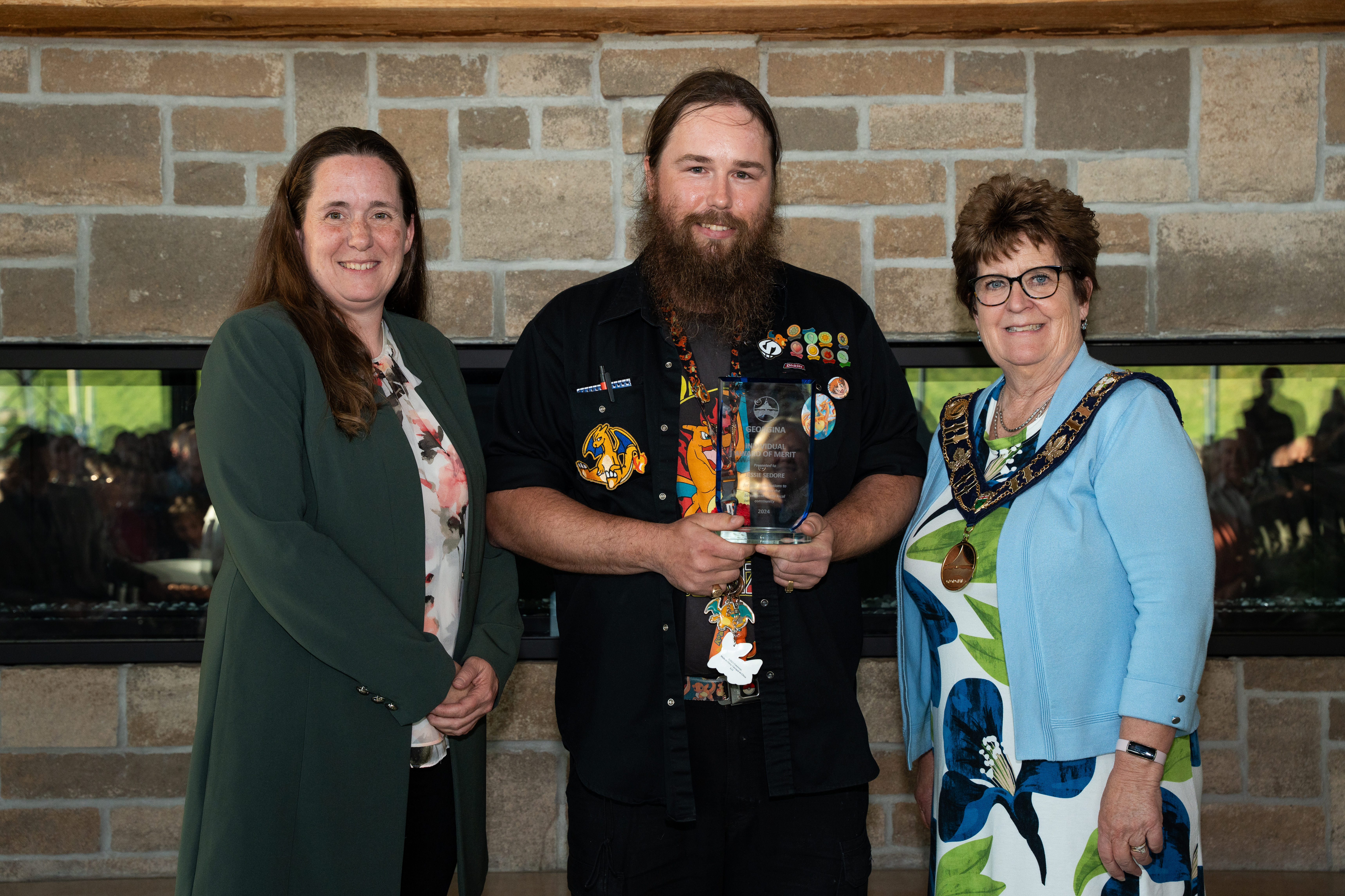 The mayor and Regional Councillor Davidson stand beside an award recipient in front of a brick fireplace, all are smiling for the photo.