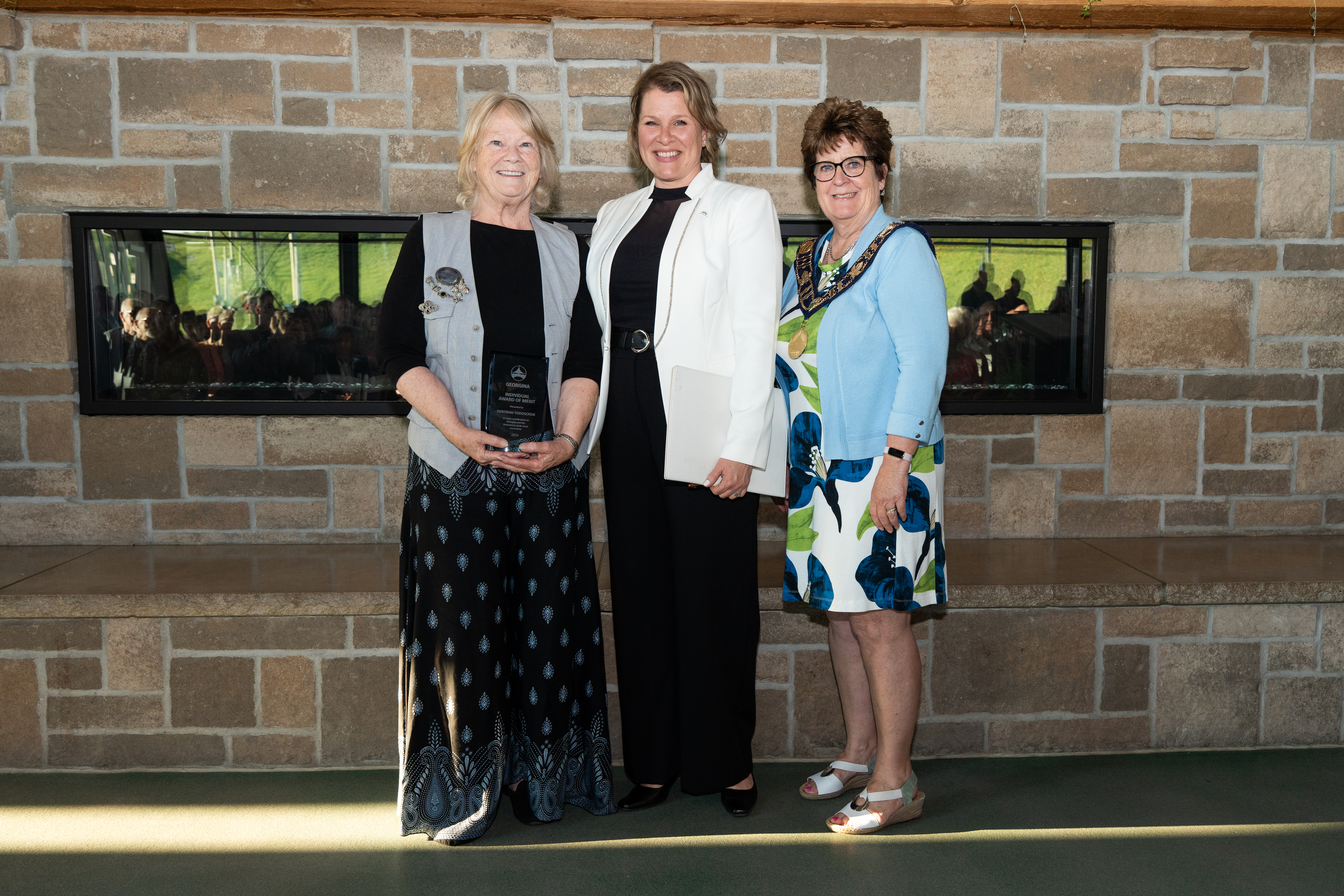 The mayor and Councillor Biggerstaff stand beside an award recipient in front of a brick fireplace, all are smiling for the photo.