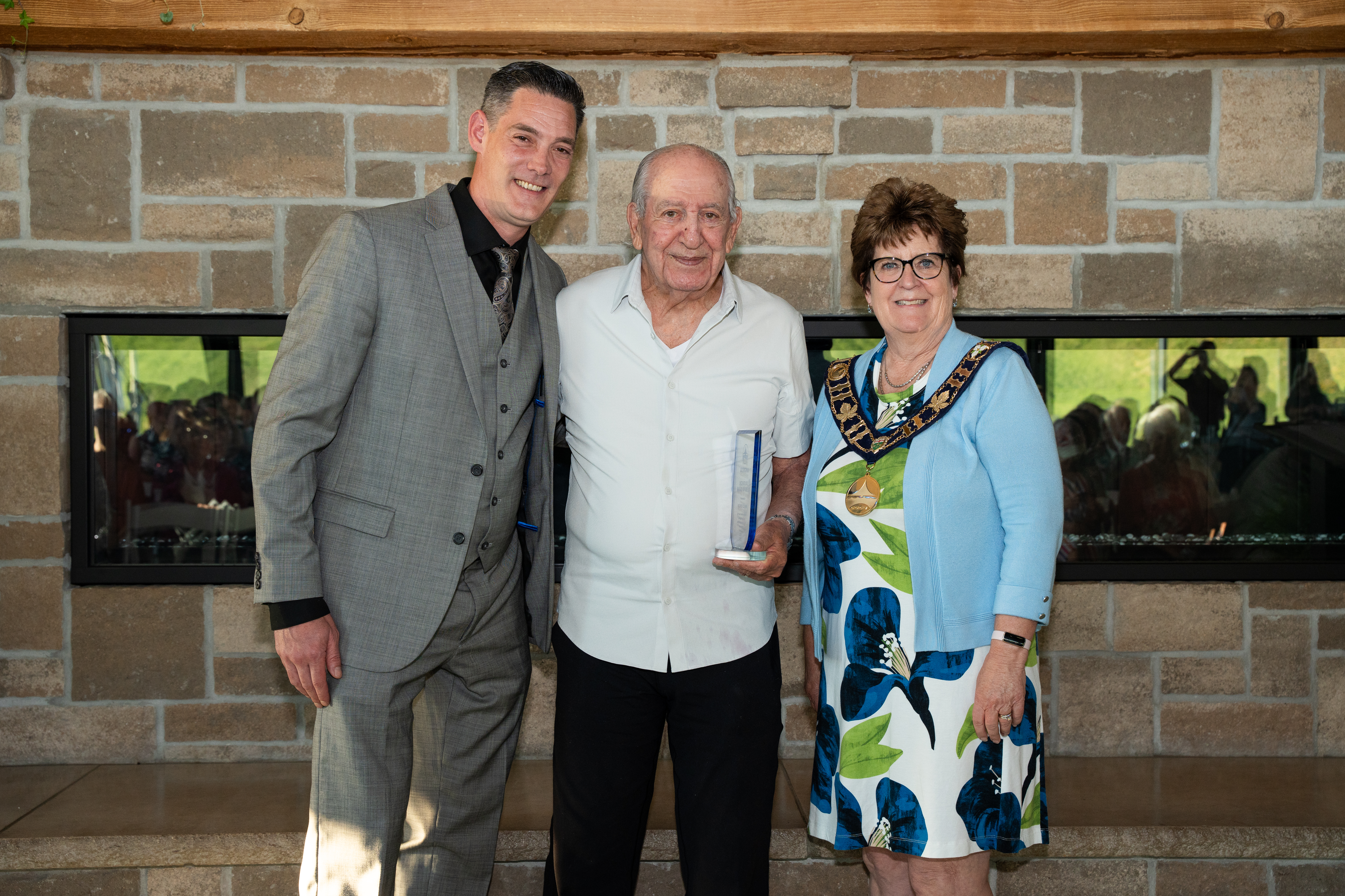 The mayor and Councillor Neeson stand beside an award recipient in front of a brick fireplace, all are smiling for the photo.