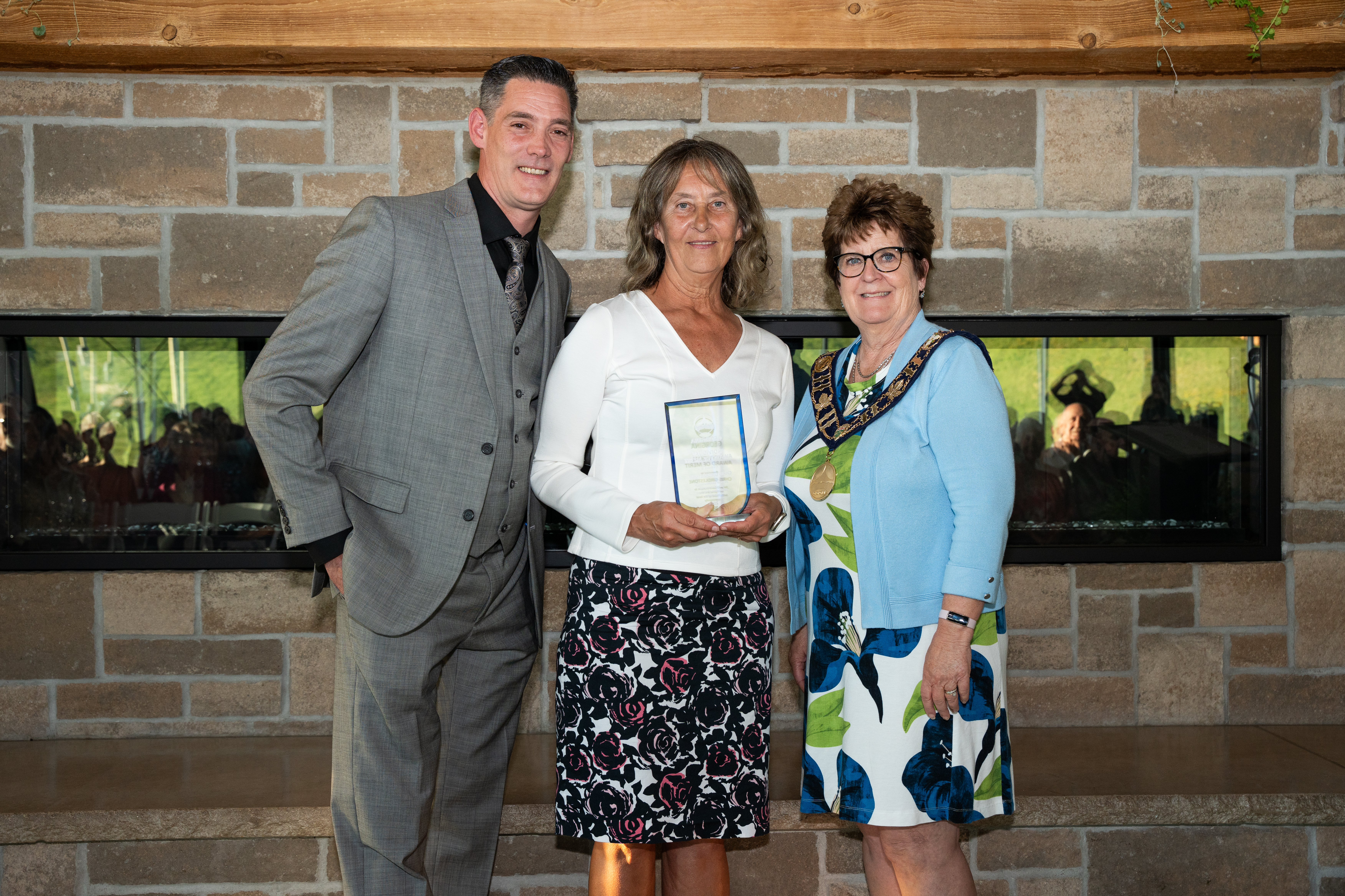 The mayor and Councillor Neeson stand beside an award recipient in front of a brick fireplace, all are smiling for the photo.