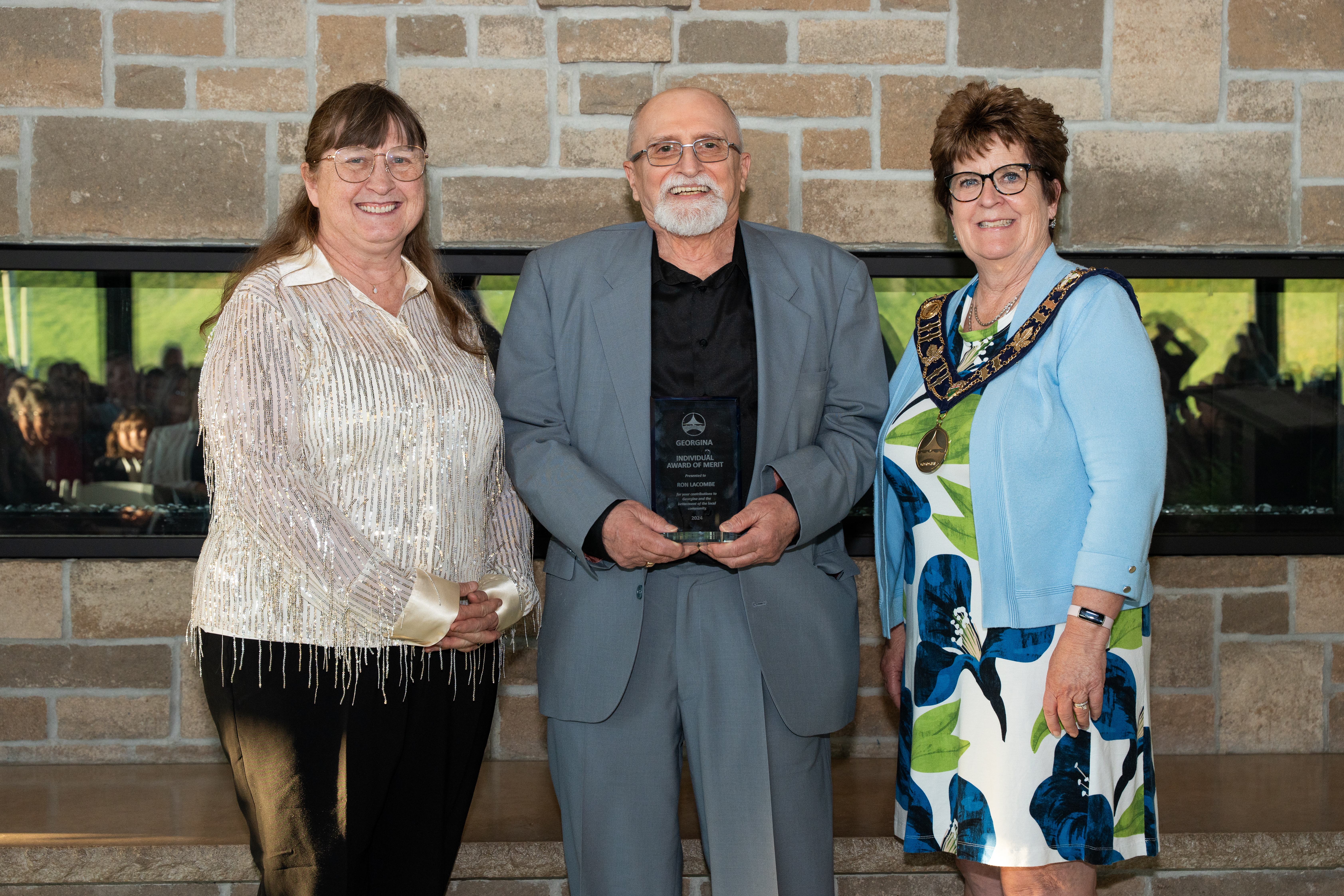 The mayor and Councillor Genge stand beside an award recipient in front of a brick fireplace, all are smiling for the photo.