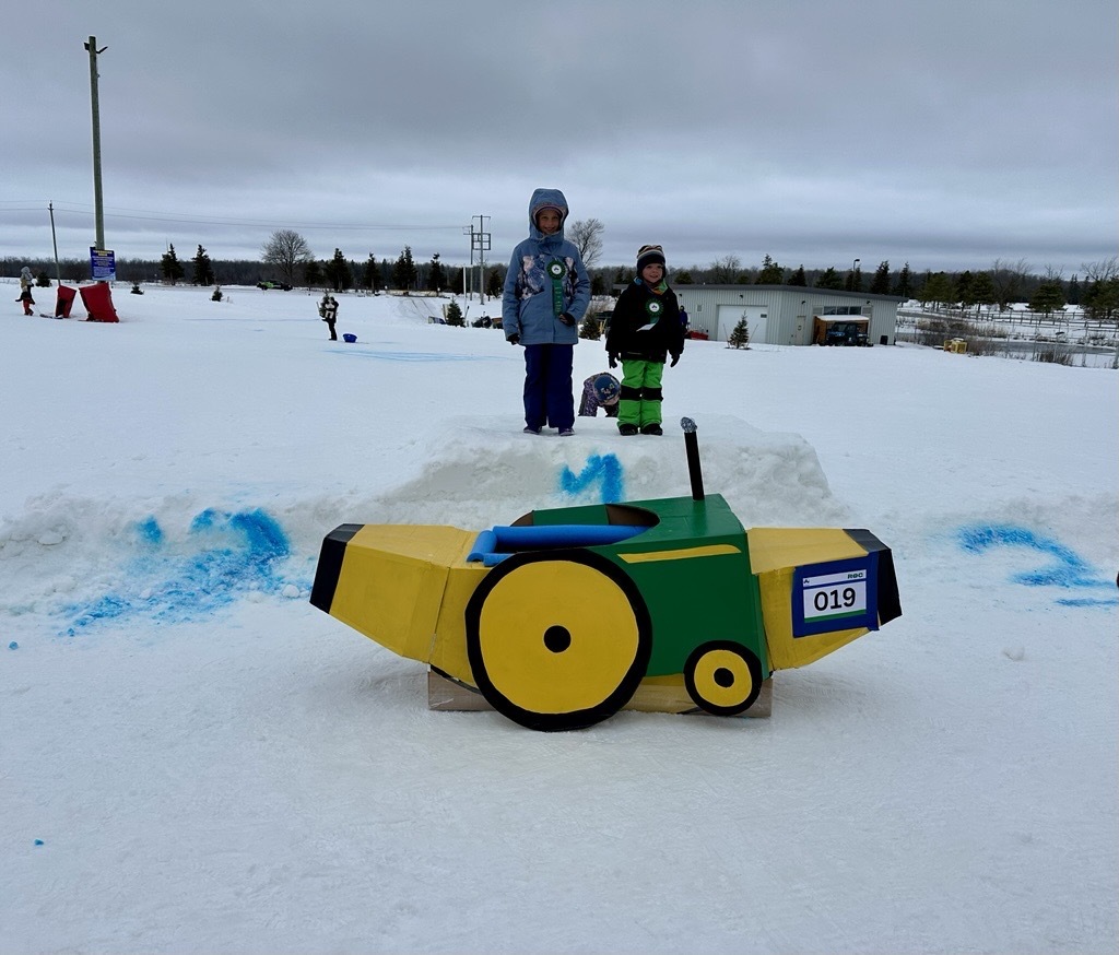 two kids standing behind a homemade cardboard toboggan that is shaped like a banana and a tractor