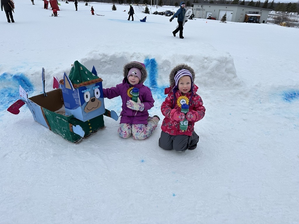 two kids kneeling next to a homemade cardboard toboggan shaped like Perry the Platypus
