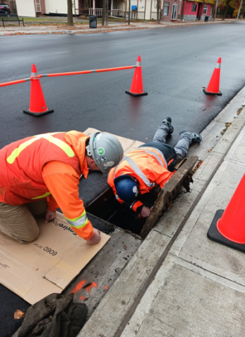 2 construction workers working on a stormwater grate with construction pylons around them