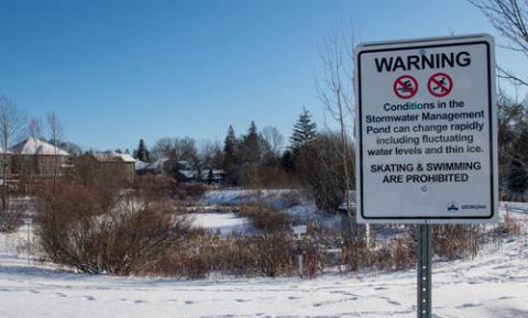 Sign warning against swimming and skating on stormwater management pond in front of a snow-covered pond in winter 