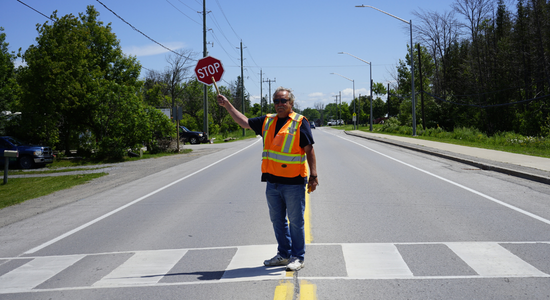 A smiling crossing guard in a high-visibility vest directing traffic on the road.