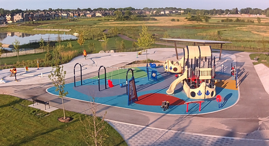 A long shot of Terry Fox Park with a wide view of the playground, green grass and a walking path under a clear blue sky.
