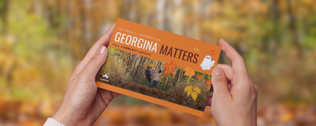 Hands holding a newsletter with the text Georgina Matters Newsletter with fall trees behind